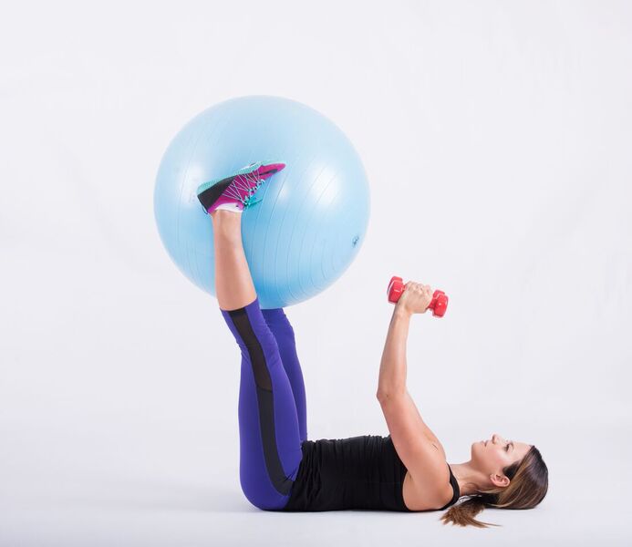 Lying on the back holding the ball between the legs with dumbbells in the hands