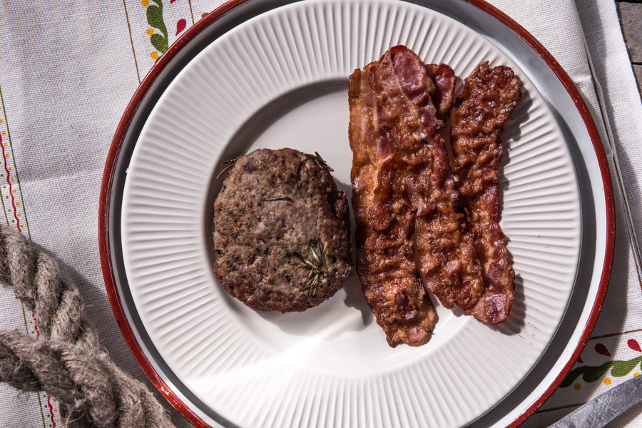 A grilled meat patty with rosemary and three rashers of crisp bacon on a white plate