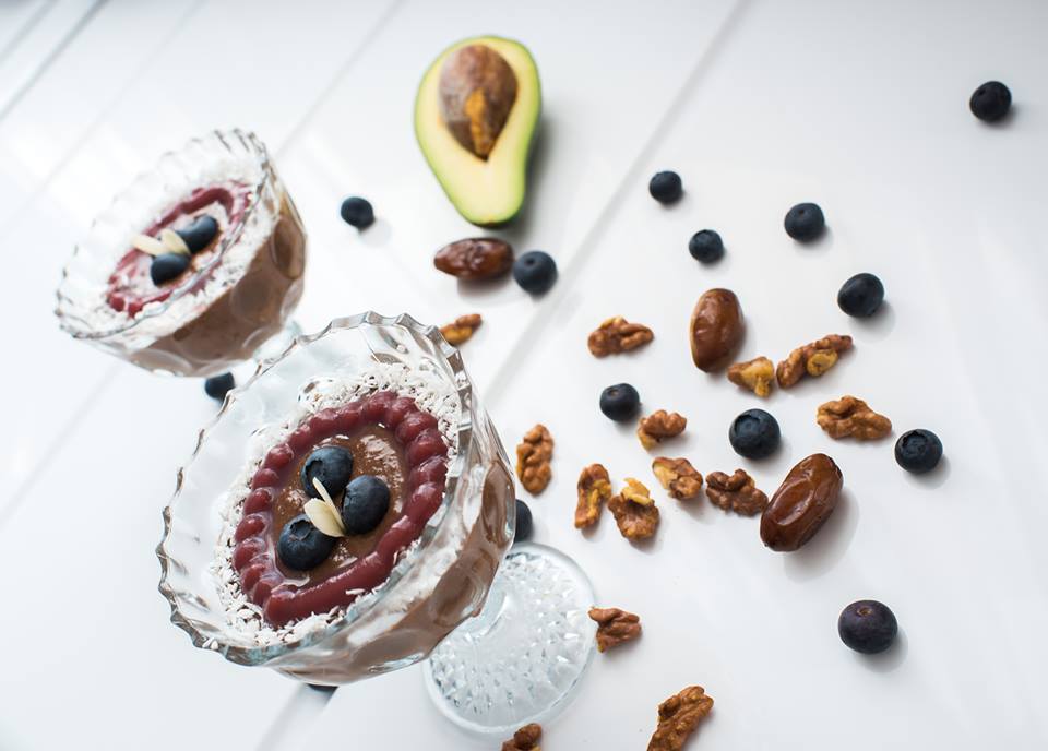 Two glass dessert cups of chocolate pudding rimmed with desiccated coconut, topped with berry glaze and blueberries, with avocado, walnuts and dates around them
