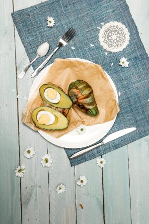 Two avocado halves filled with boiled egg and one wrapped in bacon, on a white plate