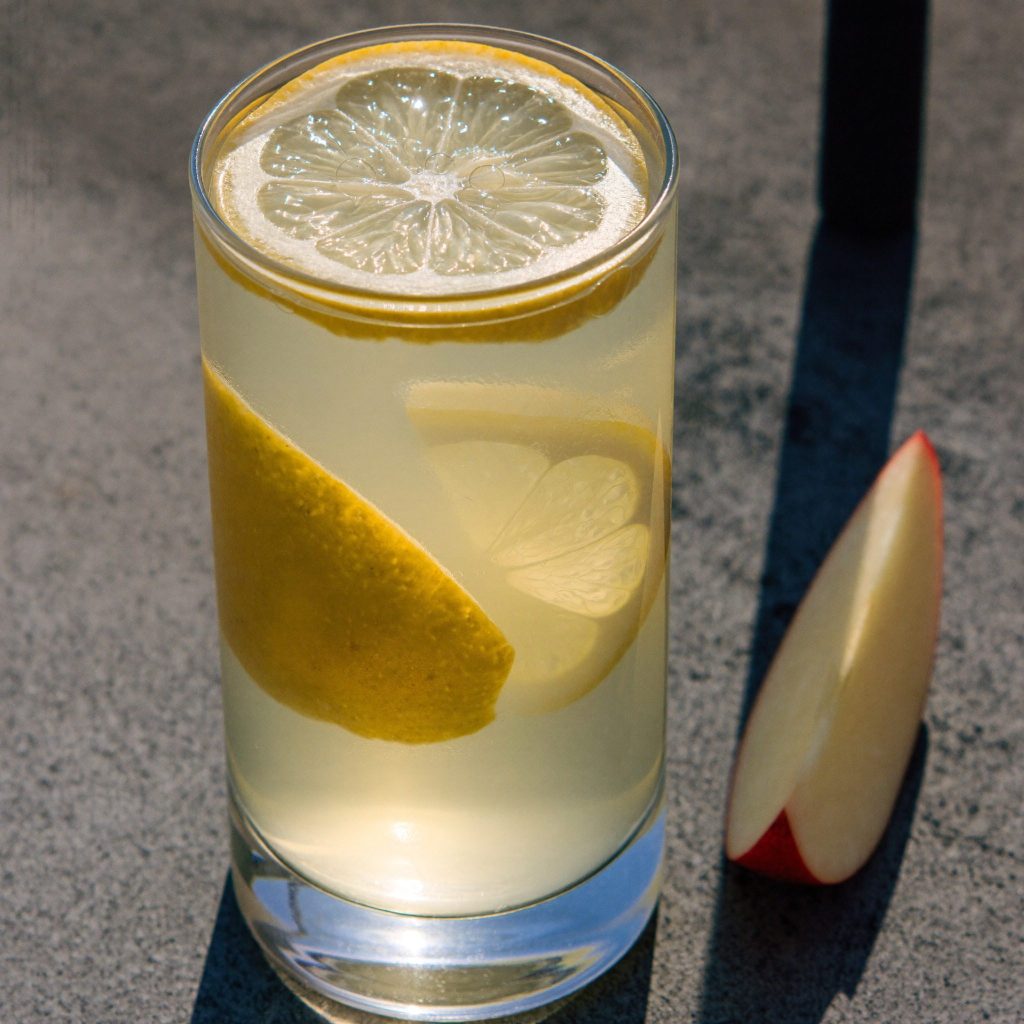A tall glass of water with lemon and apple slices, with an apple wedge beside it