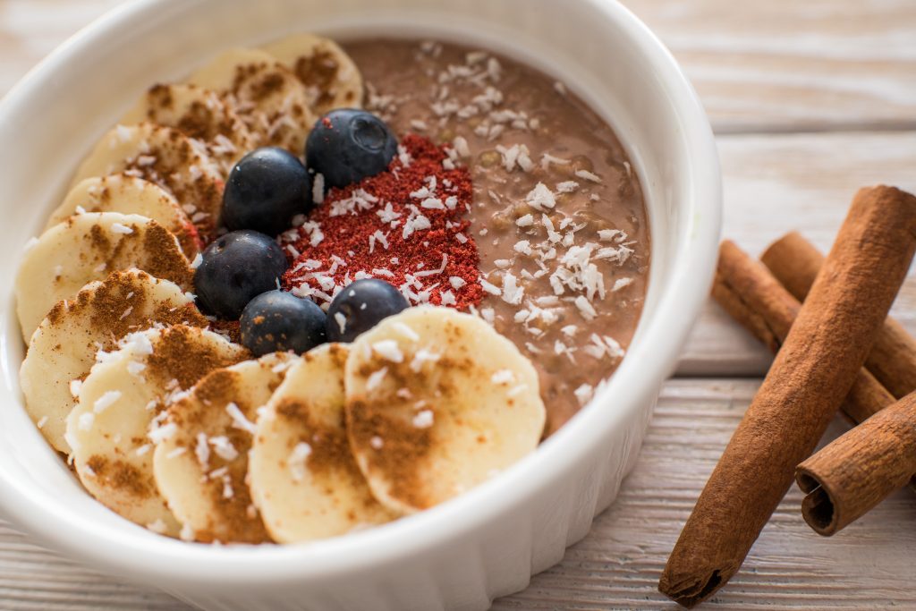 Chocolate buckwheat porridge topped with banana slices, blueberries and coconut flakes, with cinnamon sticks beside it