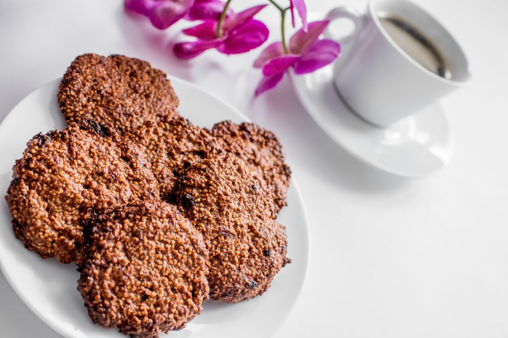 Flat amaranth cookies with raisins on a white plate, next to a cup of coffee and an orchid