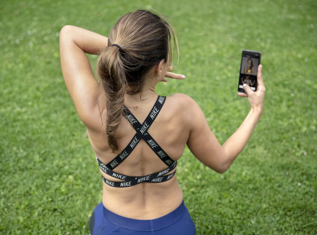 Anna Lewandowska seen from behind on grass, holding up a phone with a workout on the screen