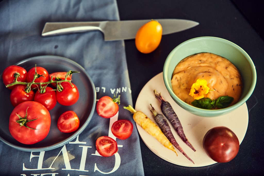Bowl of creamy sweet potato, carrot and tomato soup topped with basil and an edible flower, next to fresh tomatoes and coloured carrots