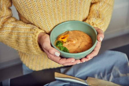 Hands in a yellow jumper holding a bowl of creamy orange soup topped with basil and an edible flower