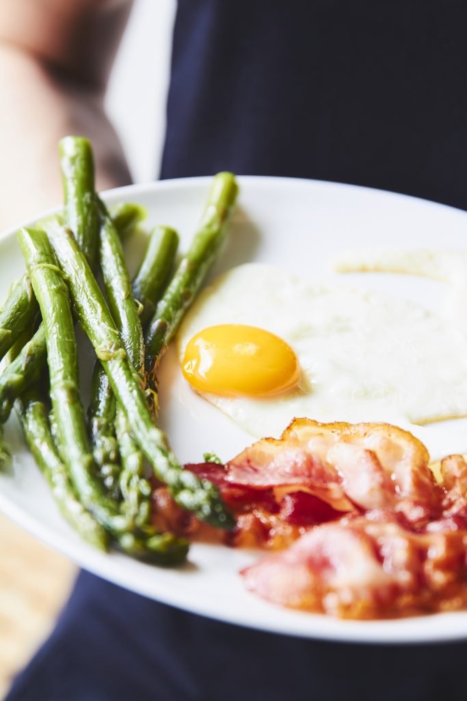 Hands holding a plate of roasted asparagus with a fried egg and bacon