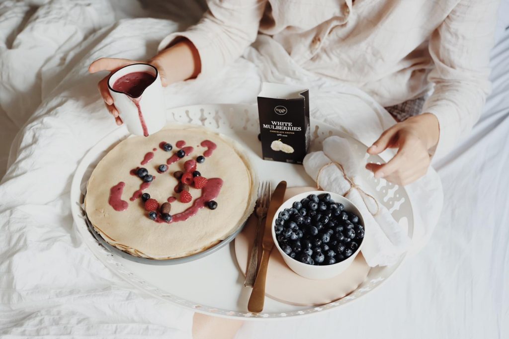 Berry sauce being poured from a jug over a stack of pancakes, with a bowl of blueberries beside it