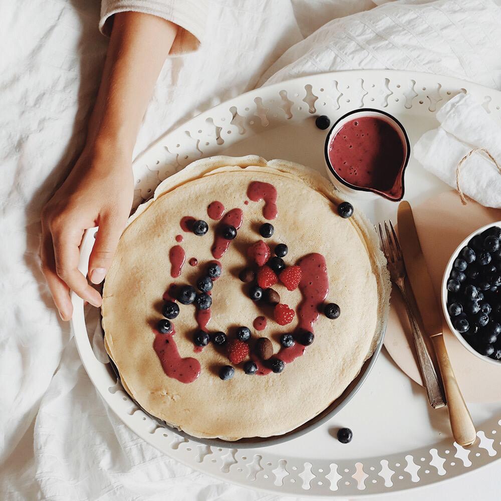 Stack of thin oat and corn flour pancakes on a white tray, topped with berry sauce, blueberries and raspberries
