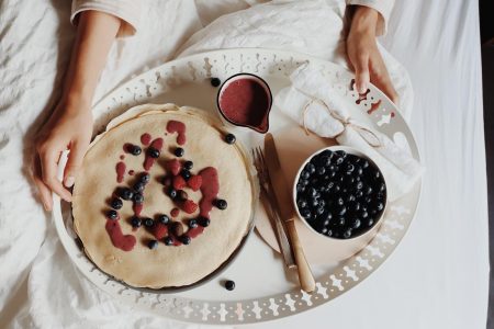 Tray with a stack of pancakes, a jug of berry sauce and a bowl of blueberries on white bedding