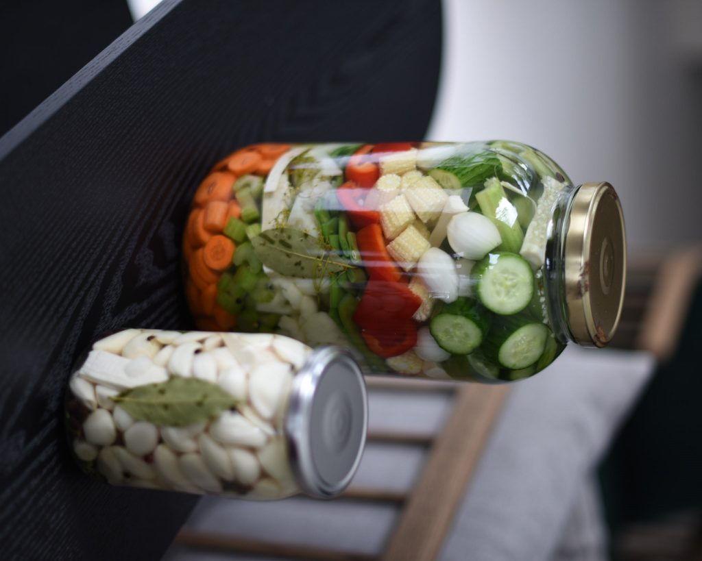 Two jars of lacto-fermented vegetables lying on a dark shelf, one with carrot, pepper and cucumber, the other with garlic