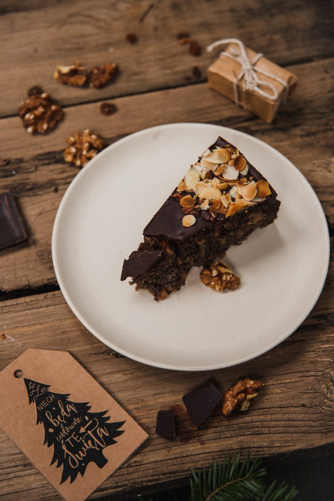 Slice of poppy seed cake with chocolate glaze and flaked almonds on a white plate, with walnuts and chocolate on a wooden table