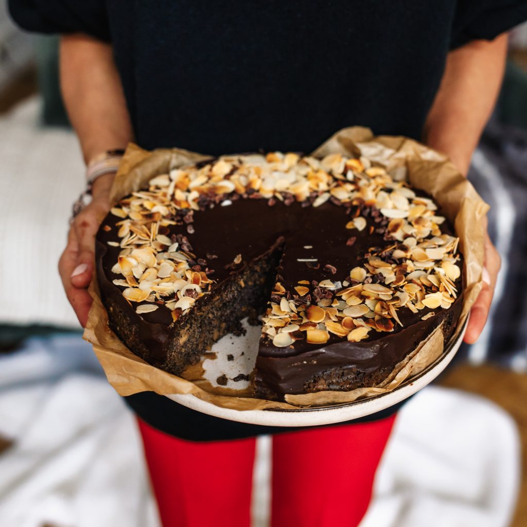 Hands holding a round poppy seed cake with chocolate glaze, flaked almonds and cacao nibs, one slice cut out