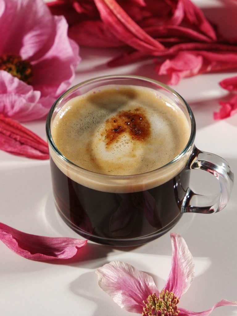 Glass cup of spiced coffee with foam and cinnamon, surrounded by pink flowers