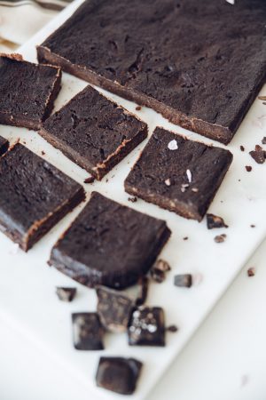 Squares of dark, fudgy brownie on a white board with pieces of dark chocolate and cacao nibs