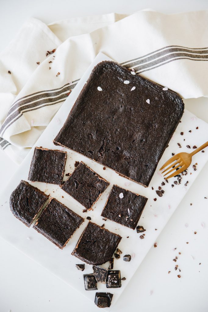 Gluten free brownie on a marble board, partly cut into squares, with cacao nibs and pieces of dark chocolate around it