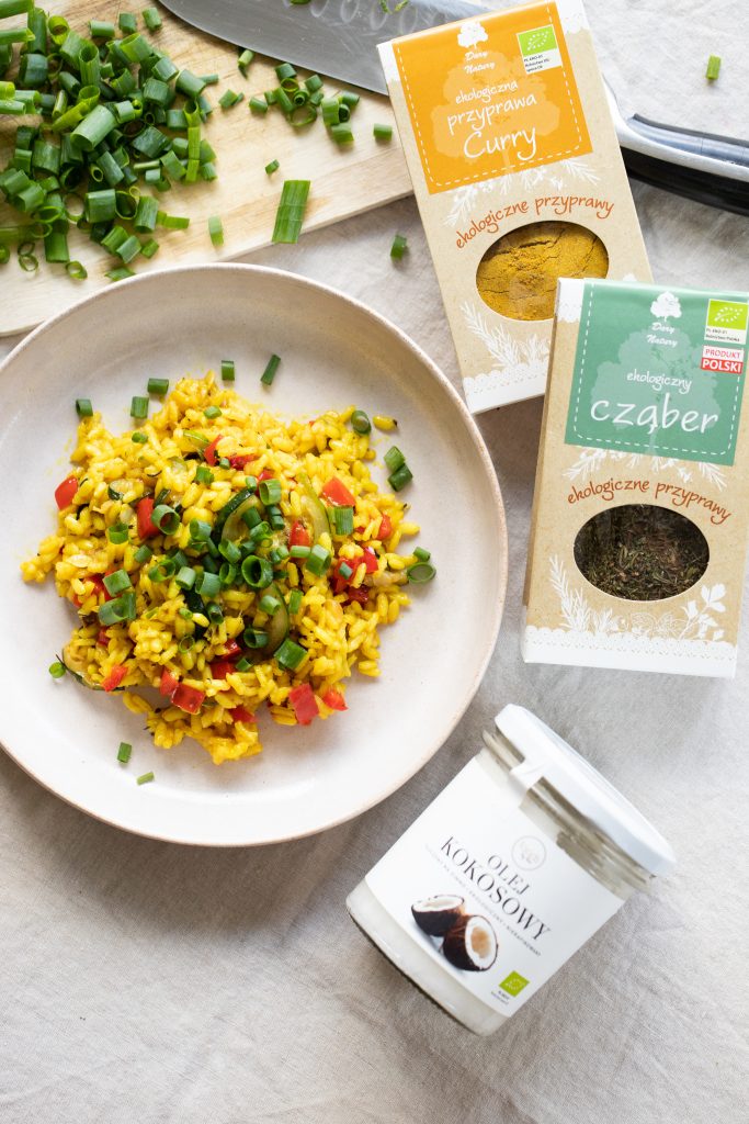 Yellow curry risotto with courgette, red pepper and chives on a plate, beside packets of curry and savory and a jar of coconut oil