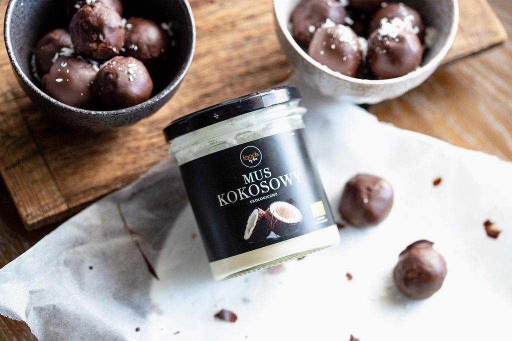 Chocolate-coated bounty balls in bowls with a jar of coconut spread on baking paper