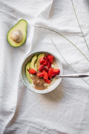 Millet porridge with avocado slices, chopped strawberries and almond spread in a ceramic bowl, seen from above