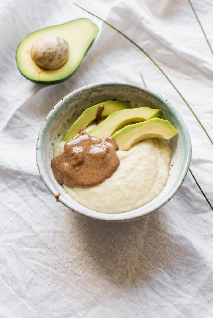 Millet porridge blended smooth in a ceramic bowl, topped with avocado slices and a spoonful of almond spread, with half an avocado beside it