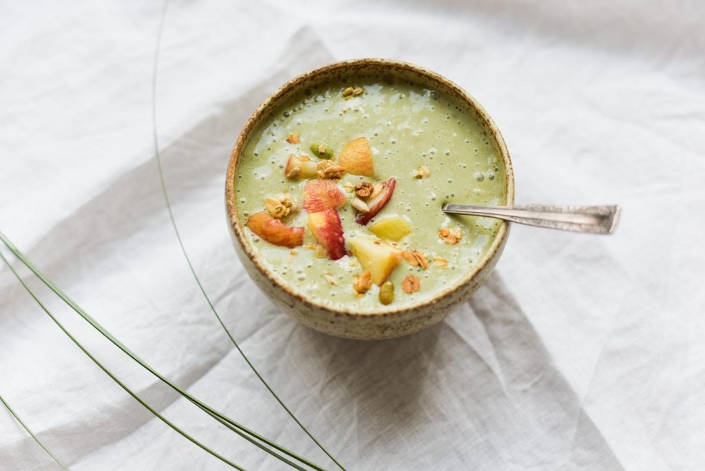 Green smoothie bowl in a ceramic bowl topped with sliced peach, apple and nuts