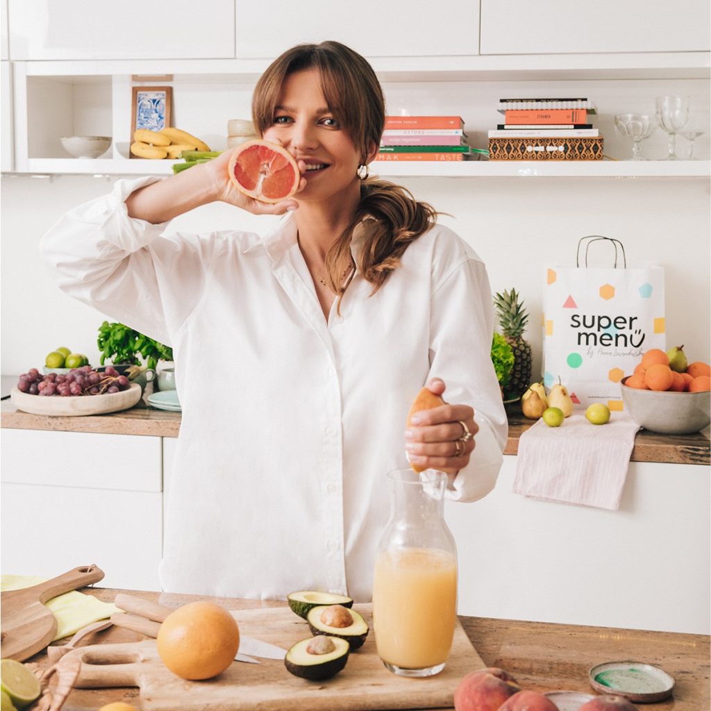 Anna Lewandowska in a bright kitchen holding half a grapefruit, with a jug of freshly squeezed juice and fruit on the counter