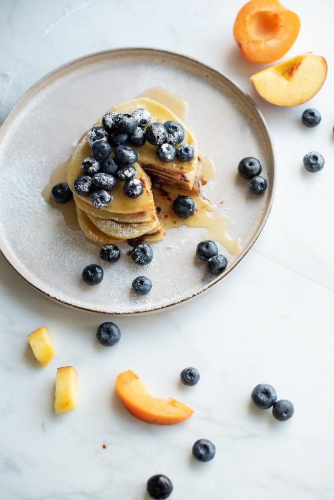 Stack of gluten-free pancakes with blueberries, honey and a dusting of xylitol, with peach slices around the plate