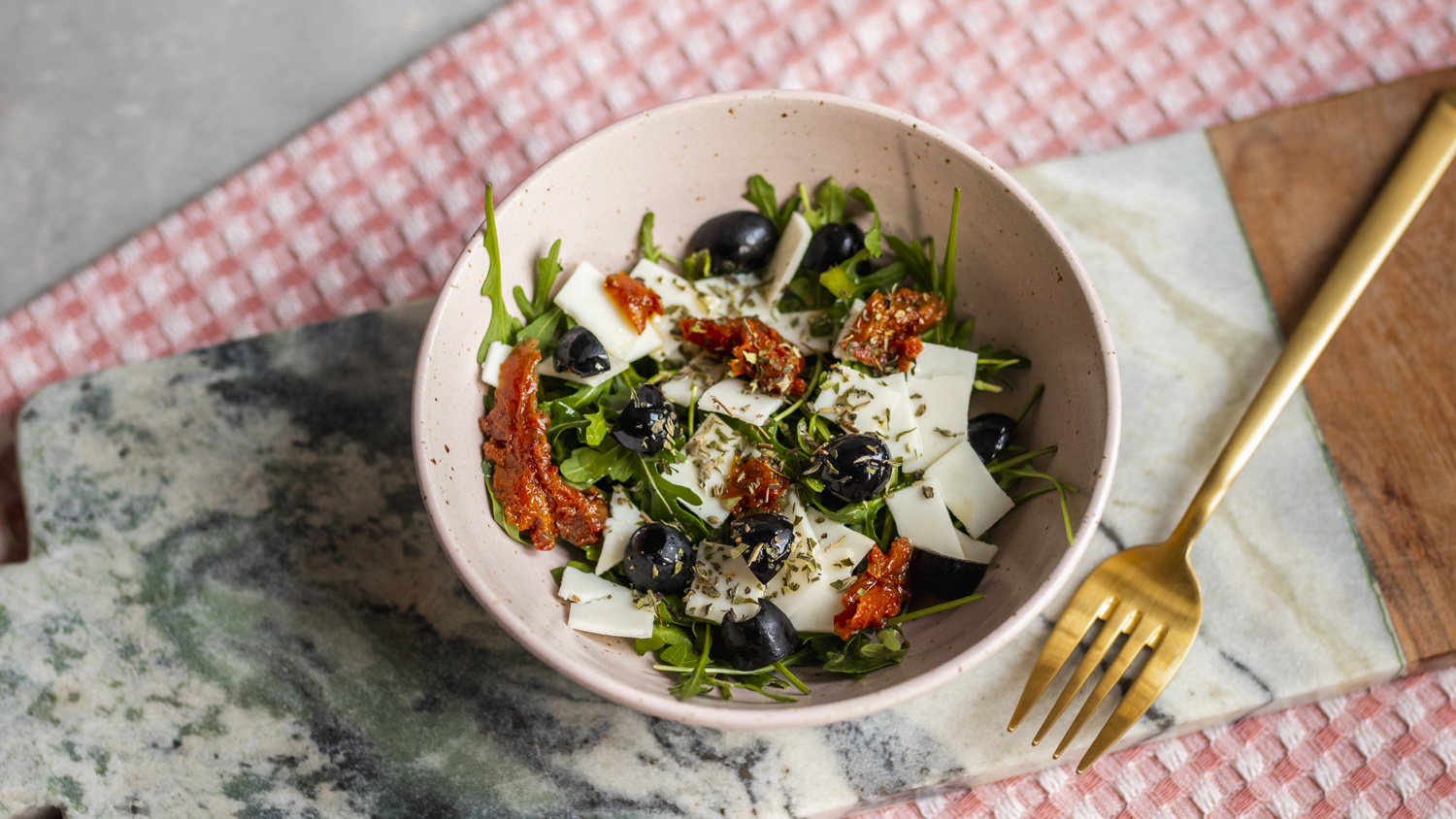 Bowl of rocket salad with black olives, strips of dried tomato and shavings of goat cheese, sprinkled with dried herbs, on a marble board