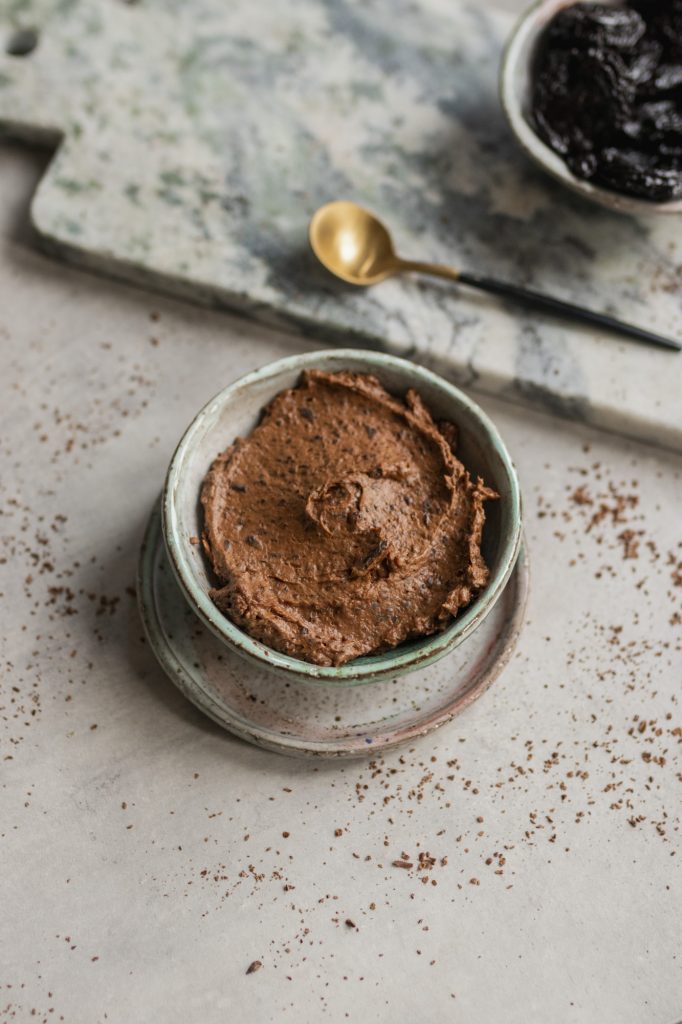Plum and chocolate mousse in a small ceramic bowl, with a gold spoon and prunes in the background