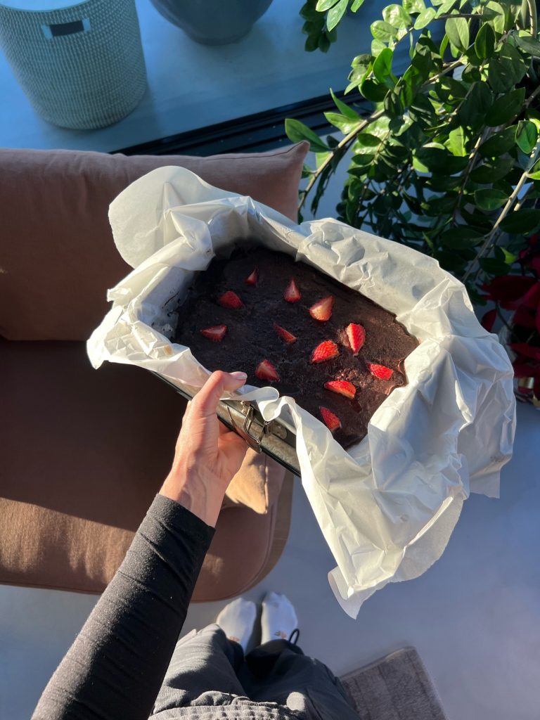 A dark chocolate brownie topped with strawberry pieces in a paper-lined tin, held in one hand