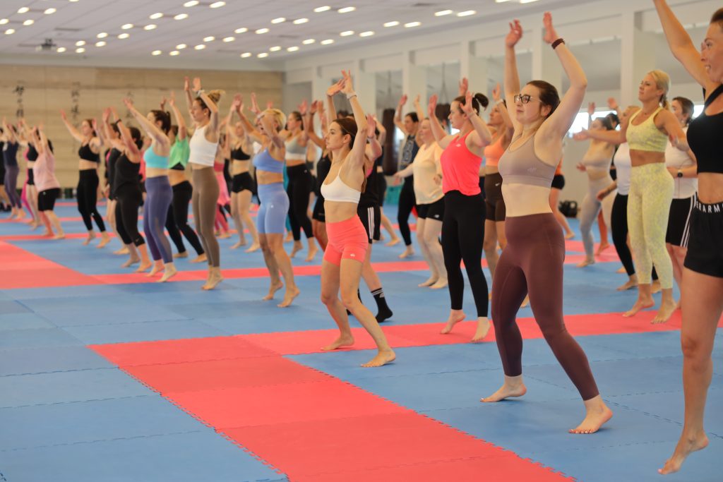 A large group training together on the mats of a dojo hall with their arms raised